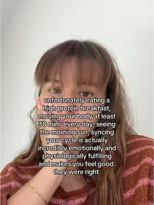 A woman with brown hair and bangs is centered in the frame against a plain white background. She appears to be looking at the camera, with her right hand touching her face near her mouth. The background is a solid, light color.  She wears a patterned sweater.
