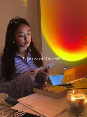 A young woman sits at a desk with a laptop, books, and a candle, illuminated by a warm, sunset-like light. Text overlayed reads 'To be successful at anything'.
