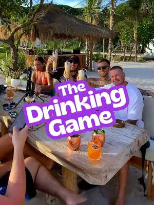 The cover image shows a group of people at an outdoor setting, likely a beach or resort, gathered around a wooden table. In the foreground, there are drinks with garnishes on the table. Overlayed on the image is a purple circular sticker with the text 'The Drinking Game' in white, bold font. The group is comprised of four people, all holding up glasses with drinks, suggesting a celebratory or social context. The overall composition is brightly lit with a focus on the people and the drinks.
