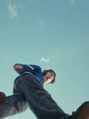 A low-angle shot of a man with headphones running or jumping, set against a clear blue sky. He wears a blue jacket and dark pants, and the word "Au" is superimposed.