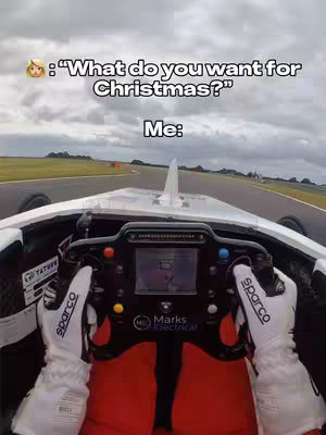 First-person perspective from inside a race car on a track. The image shows the driver's hands on the steering wheel, wearing white gloves. The car's interior is visible, with the track and cloudy sky in the background.