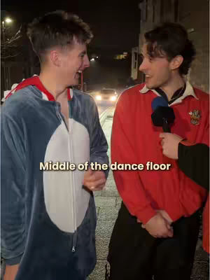 Two young men on a street at night are having an interview outdoors. One man wears a full body blue and white penguin suit, while the other is wearing a red rugby shirt and answering questions into a black microphone. The street lights and a car are visible in the background.