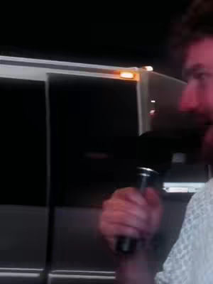 A person, holding a cylindrical object that looks like a microphone, appears to be in front of a van or large vehicle. The person is seen from the side, with focus on their face and the object they are holding.