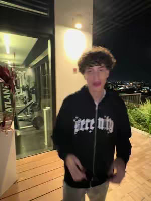 A young man in a black hoodie with white text stands outdoors at night. The background includes a glimpse of a gym, a bright overhead light, and a distant cityscape.