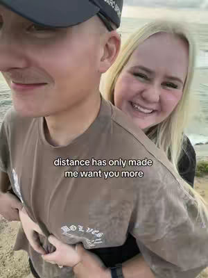 A couple is embracing outside near the water with the woman's arms wrapped around the man's waist and her head on his back. The woman is blonde, fair-skinned, and smiling. The man is fair-skinned, wearing a black hat and a brown shirt, and smiling. The text overlay reads: "distance has only made me want you more."