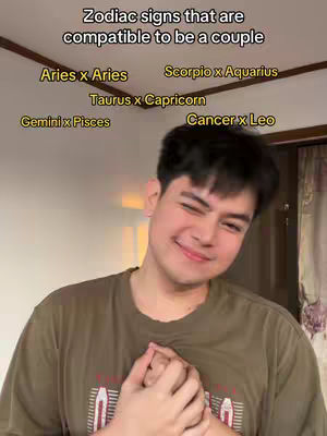A young man with dark hair and a light complexion is the central figure. He wears a casual, olive green t-shirt and smiles, winking towards the camera and clasping his hands together near his chest. Overhead, a list of zodiac signs is displayed in a white font against a somewhat blurred ceiling. The overall lighting is soft, and the background suggests a home environment.