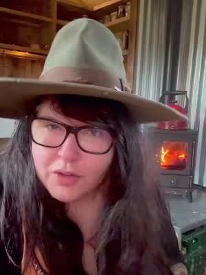 A woman with dark hair and glasses is wearing a wide-brimmed hat and looking at the viewer. Behind her is a fireplace and a wooden shelf with items on it. The image has a warm and cozy feel.