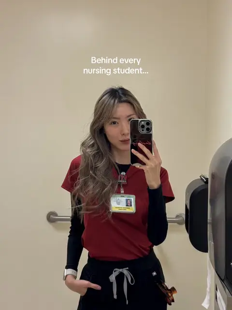 A young woman in a nursing uniform takes a mirror selfie in a bathroom. She has long, wavy hair and is holding a phone up to capture the image. The background is a plain, off-white wall with a few fixtures.