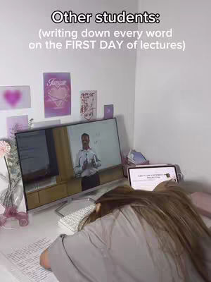 A person with long hair is lying face down on a desk with a computer, monitor and papers. The monitor has a teacher presenting a class. On a white wall in the background are posters that read: "Love"