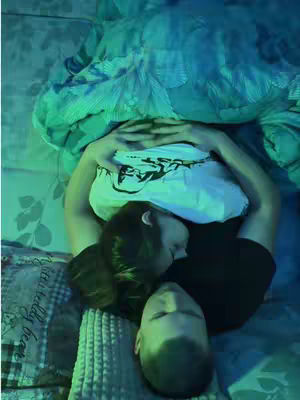 The image captures an intimate scene of a couple lying in bed, viewed from an angle above. They are nestled under a patterned comforter, with the heads of both individuals visible, suggesting a moment of closeness or relaxation. The lighting seems dim, possibly artificial, and tinted with green hues. The overall composition exudes warmth and intimacy, focusing on the connection.