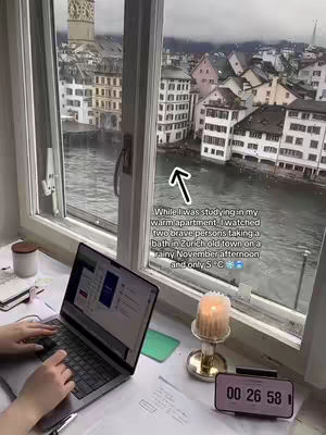 A cozy indoor scene featuring a window overlooking a cityscape with a river. A laptop sits open on a desk near the window, with a timestamp display and a lit candle nearby. The scene suggests a study or work environment with a view.