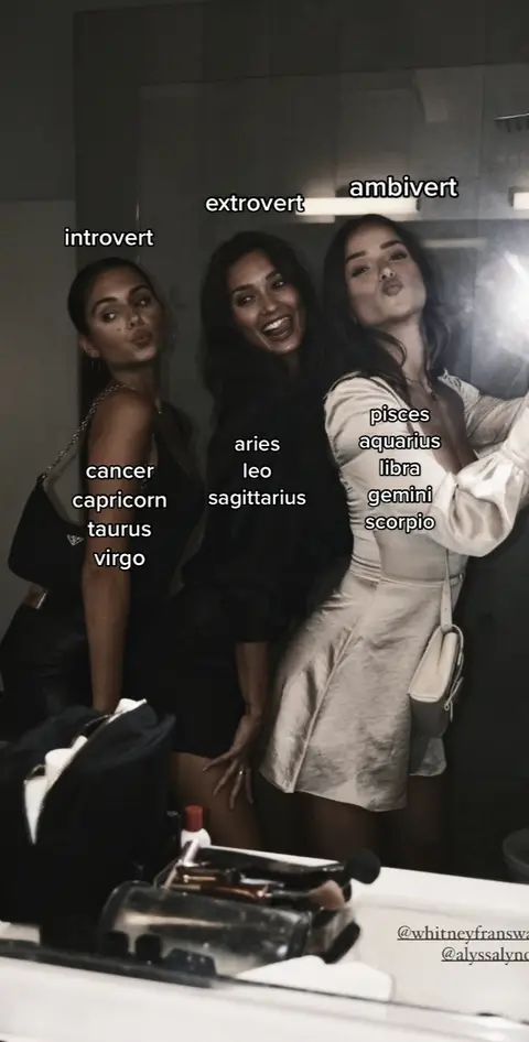 The image shows three women in a bathroom, likely taking a mirror selfie. They are positioned with astrological signs and personality labels overlaid on the mirror: "Introvert", "Extrovert", and "Ambivert". The women, dressed in dark colors, are smiling, laughing, and posing, with text labeling their zodiac signs. The setting is a well-lit bathroom, and a vanity is visible in the foreground.