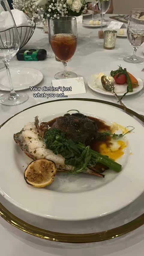 A close-up shot of a dinner plate with cooked food and some vegetables, placed on a white tablecloth. In the foreground is a plate with a piece of meat, some green vegetables, and a grilled lemon slice. The background features a blurry image of champagne glasses, silverware and an array of flowers. The text overlay reads: Your diet isn't just what you eat...