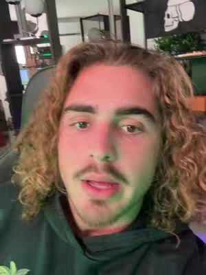A man with long, curly brown hair and a mustache is centered in a selfie-style shot. He's looking at the camera and talking, wearing a dark hoodie. The background includes glimpses of an office setup with computer monitors and other items.