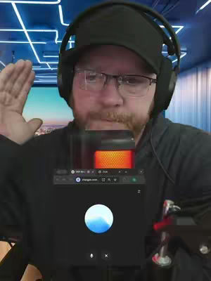 A man wearing a black cap, glasses, and headphones is looking toward the camera. He has a beard and is holding up his right hand. Behind him is a blurred background with a neon blue design. In front of him, he's holding a microphone that has a small screen with a webpage or app running. The overall composition is a selfie style with the man in the center.