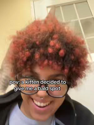 A selfie-style image featuring a person smiling broadly. The person has red curly hair, glasses, and a nose ring. The text overlay reads: "pov: a kitten decided to give me a bald spot". The background includes a tan wall and a white door with windows.