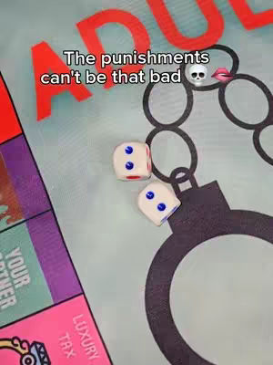 The image is a close-up of a game board, likely a couples' board game. Two dice are visible, showing a roll of 5 and 2. Above the dice, there is text that says "The punishments can't be that bad", along with a skull and a pair of red lips. A large handcuff graphic is prominent. The overall tone suggests playful challenges and risqué themes.