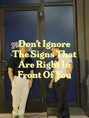 The image shows two men standing in front of a door. Text overlayed on the image says 'Don't Ignore The Signs That Are Right In Front Of You'. One man is wearing a black shirt and white pants, and the other man is wearing a white shirt and black pants.