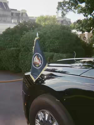 The cover image showcases a black car with a presidential flag on the front. The flag, black with a circular emblem, is attached to the car's front. The car's reflective surface and wheel are also visible. A building with sunlight and green foliage are in the background.