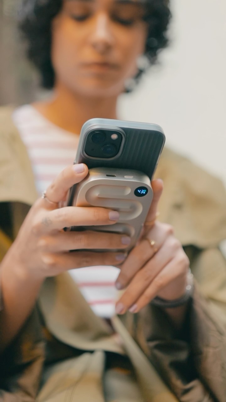 A medium shot focusing on a woman who is holding her phone with a power bank attached. The phone is in focus with the camera of the phone clearly seen, as well the top of the power bank. The woman's face is slightly blurred, but she appears to be looking down at the phone. She is wearing a light-colored striped shirt underneath a green coat. Her nails are painted with a very pale pink nail polish. The background is mostly out of focus, but appears to be a neutral color.