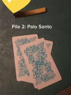 The image shows three tarot cards laid out on a dark green surface. A piece of palo santo wood sits above the cards. A yellow shape with a star in it is in the top left corner. Text overlays read "Pile 2: Palo Santo".