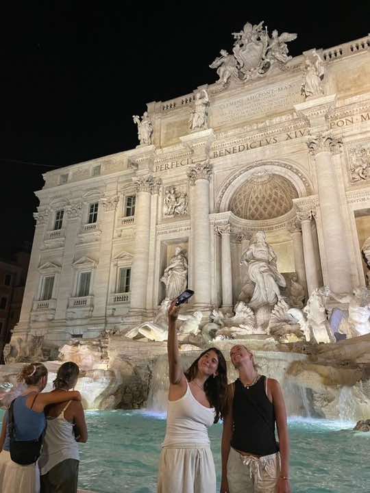 Two young women taking a selfie in front of the Trevi Fountain at night. The fountain is illuminated, and other people are visible nearby.