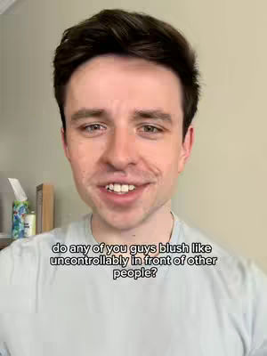 A man with short dark hair and a light blue t-shirt is centered in the frame. The background is a light beige color. A text overlay at the bottom of the frame asks, "do any of you guys blush like uncontrollably in front of other people?"