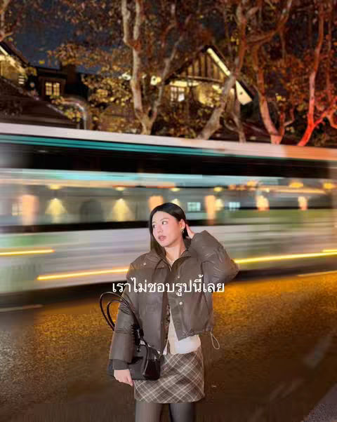 A woman stands on a city street at night, in front of a blurred background of a train and buildings with lights. She is wearing a brown jacket, a skirt, and holding a purse.