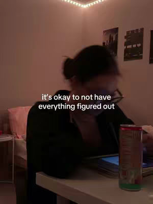 A dimly lit room with a woman sitting at a desk with an open laptop and a drink. The text overlay reads: 'it's okay to not have everything figured out'. The background features a bed with a pink pillow, some artwork, and a string of lights.