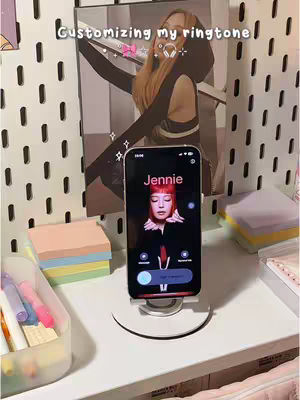 A phone displaying an incoming call from Jennie. The phone is on a white stand on a white surface. Behind the phone is a board with a picture of Jennie and other items.