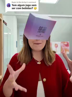 A woman in a red coat and a white top is holding a box in front of her face, hiding most of it. She's in a white-walled room with some objects in the background. Her hands are up, with fingers spread, as if she's about to grab something.