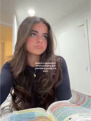 A young woman with long brown hair looks upward pensively, sitting indoors. She's wearing a dark shirt and a delicate gold necklace. In front of her is an open book with colorful pages. The background is slightly blurred, but appears to be a room with neutral-colored walls and a light fixture.