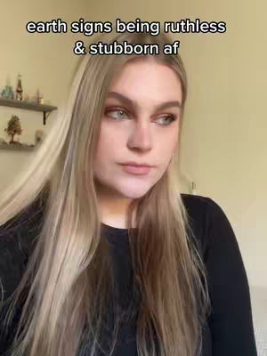 A blonde woman with long hair and a black long sleeve shirt is looking off to the side, with a neutral expression on her face. The background is a room with a cream-colored wall and a shelf with decorative items