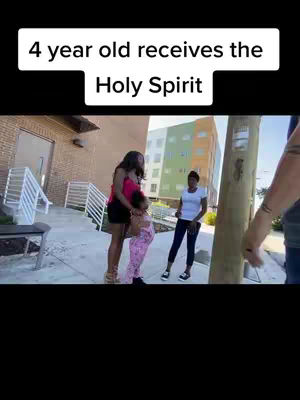 The image shows a young black girl being held by an adult black woman, possibly her mother, who is standing in front of a building with other people in the background.