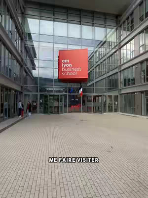 A wide shot of a modern building facade with a large red square sign that reads 'emlyon business school'. The foreground shows a patterned brick pavement. Below, a text overlay reads 'ME FAIRE VISITER'. The architecture is a mix of glass and metal, reflecting the sky. The angle points towards the main entrance of the building.