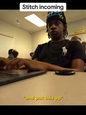 A young Black man sits at a desk in a classroom, typing on a laptop. He wears a black shirt with a white logo and a patterned beanie. The text at the top says "Stitch incoming" in a white box, and a caption at the bottom says, "and put this up". The background shows other students seated, and a yellow sign on the wall