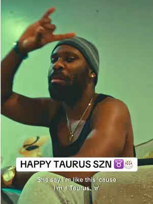 A man is featured, positioned slightly off-center, gesturing with his right hand. The background shows a light green wall. The man is wearing a beanie, a black tank top, a watch and a necklace. Text overlays include 'HAPPY TAURUS SZN' and a caption about being a Taurus. The overall composition is casual.