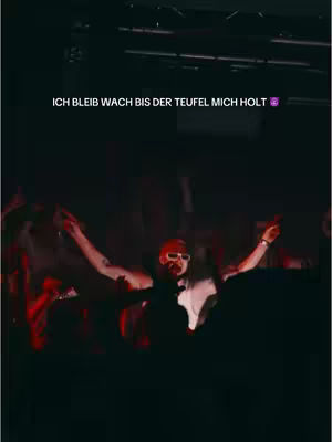A dark, concert-like scene with a performer at the center, arms outstretched. The foreground is filled with silhouettes of people. The background is dark with stage lighting.