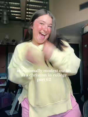 A cheerful young woman with long brown hair and rosy cheeks is the central figure. She is smiling widely with her mouth open and is wearing a light yellow long-sleeved shirt over pink pants. The background features a window with blinds and some interior elements, including what appears to be a closet or cabinet.
