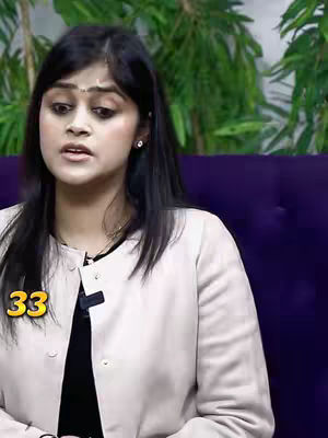 A woman is centered in a medium shot, slightly angled to her left, with a soft focus. She has dark hair, long and flowing, with subtle waves. She wears light makeup and has a small dot on her forehead, a traditional Indian decoration. She's wearing a light cream-colored jacket over a black shirt. In the bottom left corner, the number "33" in a yellow sans-serif font is overlaid. The background includes green plants with a blurry appearance and a purple surface to the right of the woman.