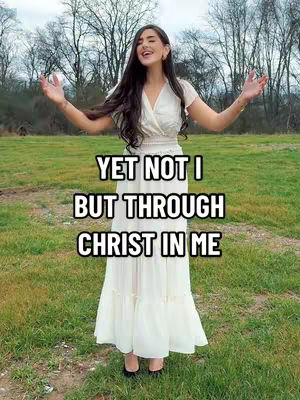 A woman with long dark hair is standing in a green field with her arms outstretched in front of a blurry backdrop of trees under a blue sky. Centered over her is text in a stylized font.