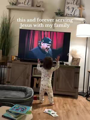 A living room scene with a child in the foreground and a television playing a video in the background. The child, wearing white pajamas, has their arms raised in the air, seemingly mimicking the person on the television screen. The room has a neutral color palette.
