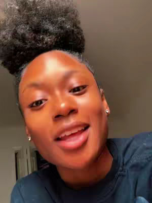 A close-up shot of a Black woman with her hair in a bun, smiling and looking towards the camera. She is wearing a dark colored shirt, and there are small earrings visible. The background is out of focus but warm-toned.