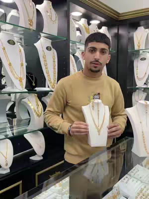A man stands behind a glass counter holding a display piece with a gold necklace. Behind him, a storefront features more jewelry displays, creating a glamorous setting. The man is wearing a light brown sweater. The necklaces on display are gold with elaborate patterns.