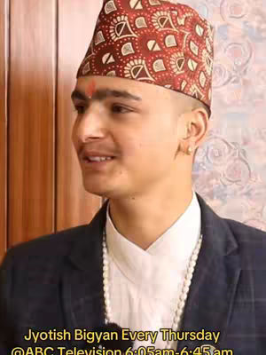 A medium shot of a young man in a traditional Nepali hat and outfit, smiling as he is posed. He has a red mark on his forehead and a necklace. The background is softly blurred with neutral colors.