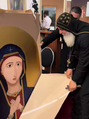 The cover image shows a man, presumably a bishop, and another person unveiling an Orthodox Christian icon. The icon depicts the Virgin Mary, or Theotokos, with a blue veil. The bishop, dressed in black robes with a cruciform pectoral cross, is holding the cover of the icon with both hands, while another person wearing a white shirt and black suit is helping him. The background includes parts of a wooden table. There are some additional elements, like cameras and possibly another person in the background.