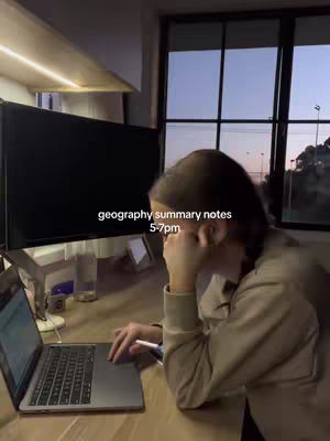 A person is sitting at a desk with a laptop, appearing to study late at night. The room is dimly lit, with a large computer screen in the background and a window showing a twilight sky. Text overlayed indicates geography summary notes being studied between 5-7 pm.
