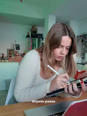 A young woman with long brown hair is seated at a wooden desk, concentrating on writing with a stylus on a tablet. She wears a white long-sleeved shirt. The room is brightly lit with a mint green wall, and there's a laptop open next to her. A sign reading 'ATING' and some boxes are visible, alongside some wall hanging items and green boards in the background.