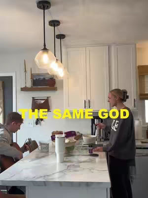 A well-lit indoor scene, likely a kitchen, features two people. A man is seated on the left playing a guitar, with the instrument visible. A woman, standing to the right, is in the midst of a gesture with her hands. The room has white cabinets and countertops, with warm lighting from pendant lights above. A text overlay in yellow with the phrase 'THE SAME GOD' is present in the upper center. The overall composition suggests a casual, domestic setting.