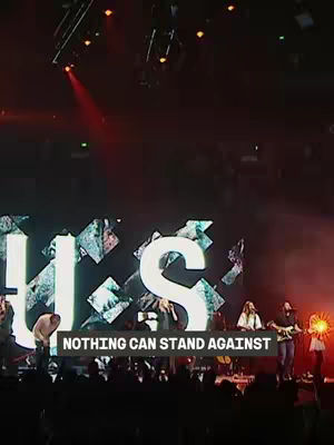 A live performance on a stage with large illuminated letters in the background. The foreground features performers and equipment. A text overlay at the bottom reads a caption. Overall the lighting is moody, with red dominating.